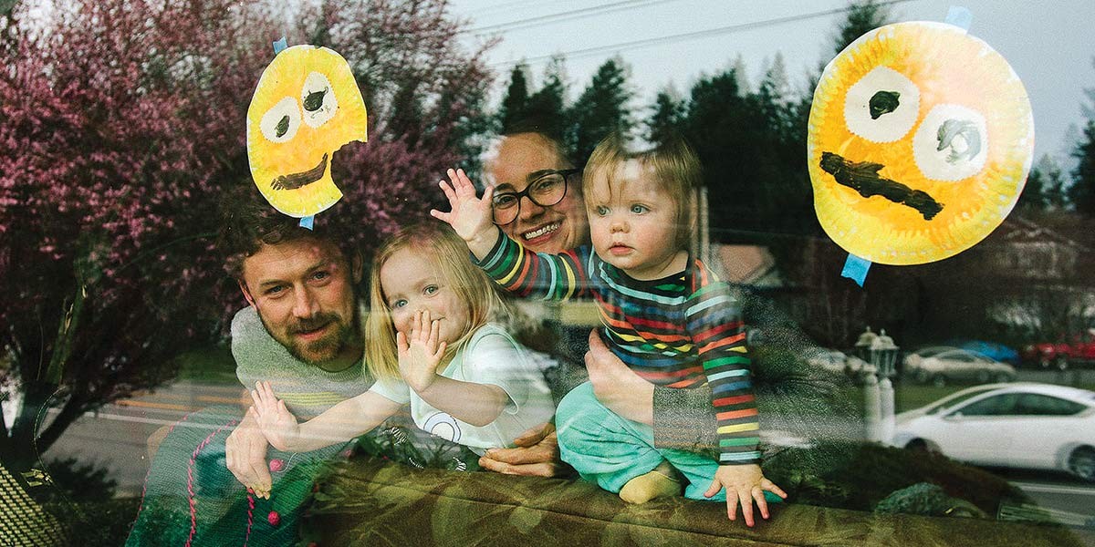 Family with two children waving through a window decorated with handmade yellow smiley faces.