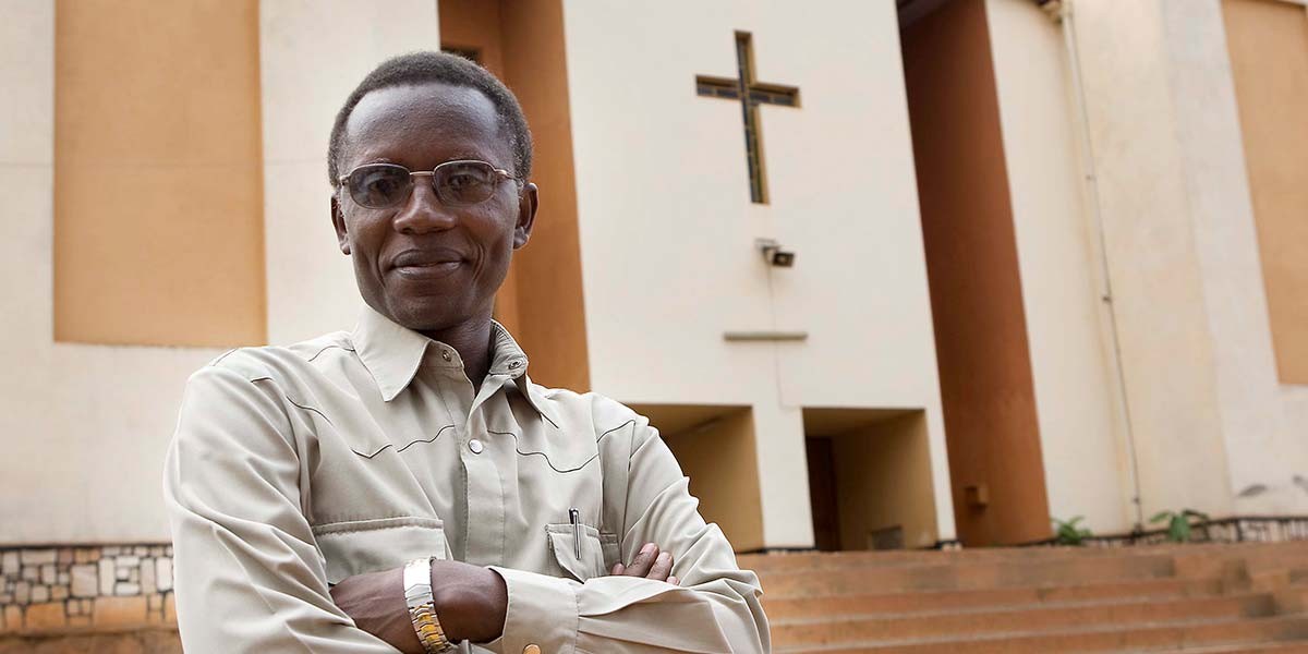 Man with glasses and beige shirt standing with arms crossed in front of a building with a cross.