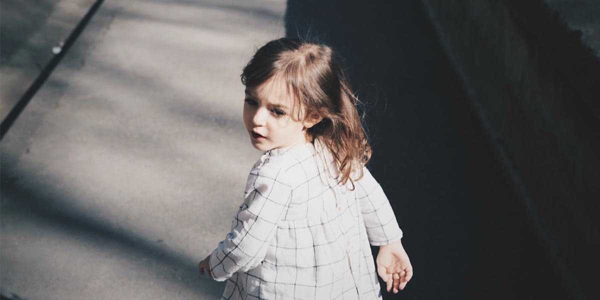 Young girl in a white checkered dress looking back while standing on a sunlit concrete path.