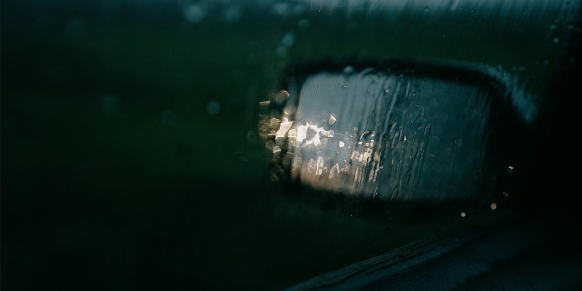Raindrops on a car side mirror reflecting blurry light during a dark rainy scene.