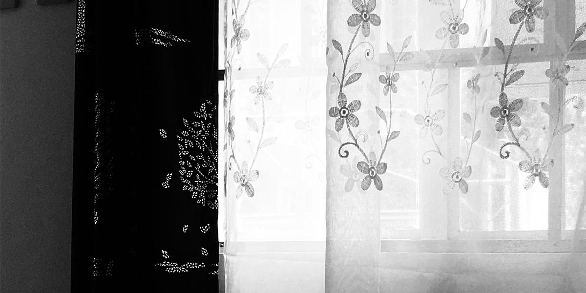 Window with sheer white floral curtains beside partially visible dark patterned curtain.