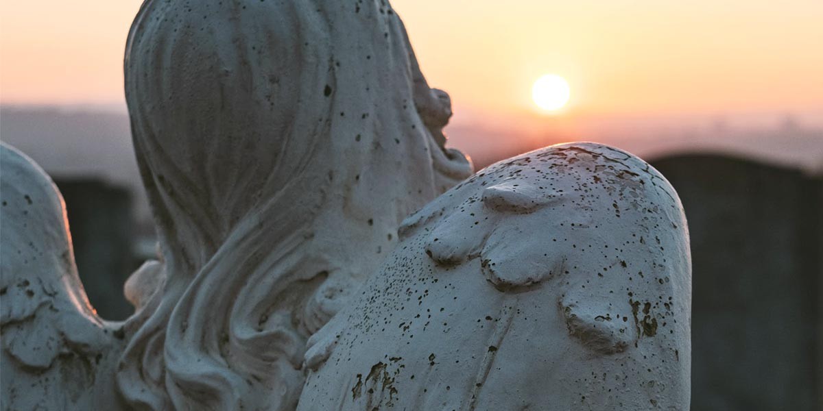 Close-up of a weathered angel statue's head and wings against a sunset sky.