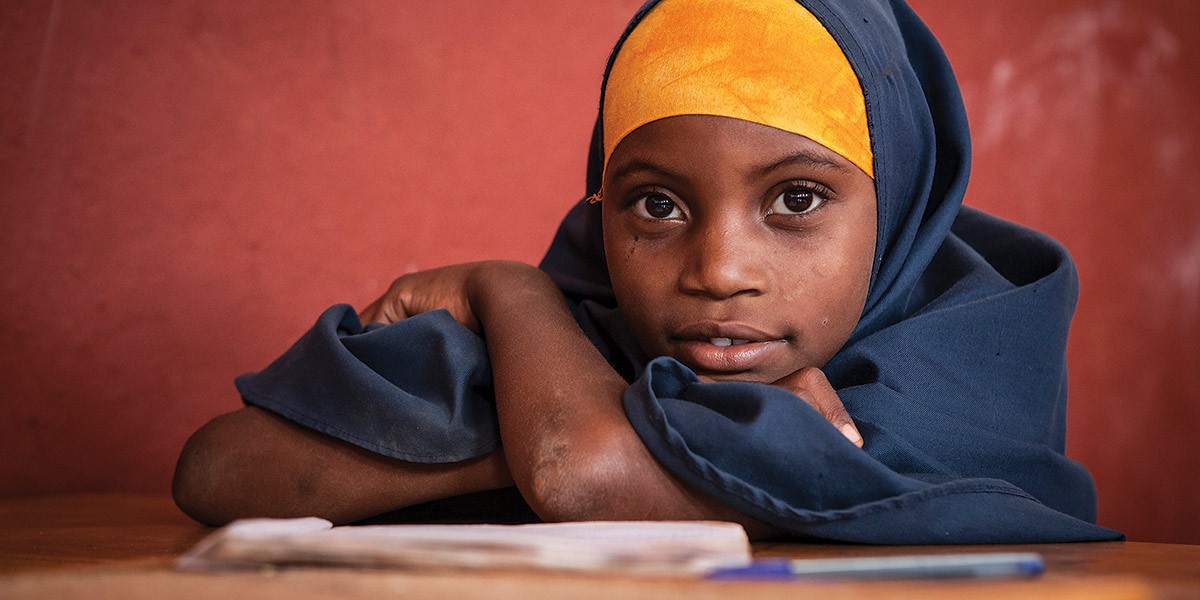 Young girl wearing a blue hijab with a yellow headband resting on her arms at a desk with a pen and notebook.