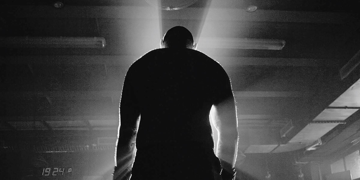 Backlit silhouette of a person standing in a dimly lit industrial or gym space with light rays behind.