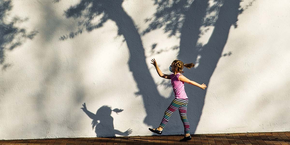 Girl wearing a mask and colorful striped pants walks by wall with a tree shadow and her shadow.