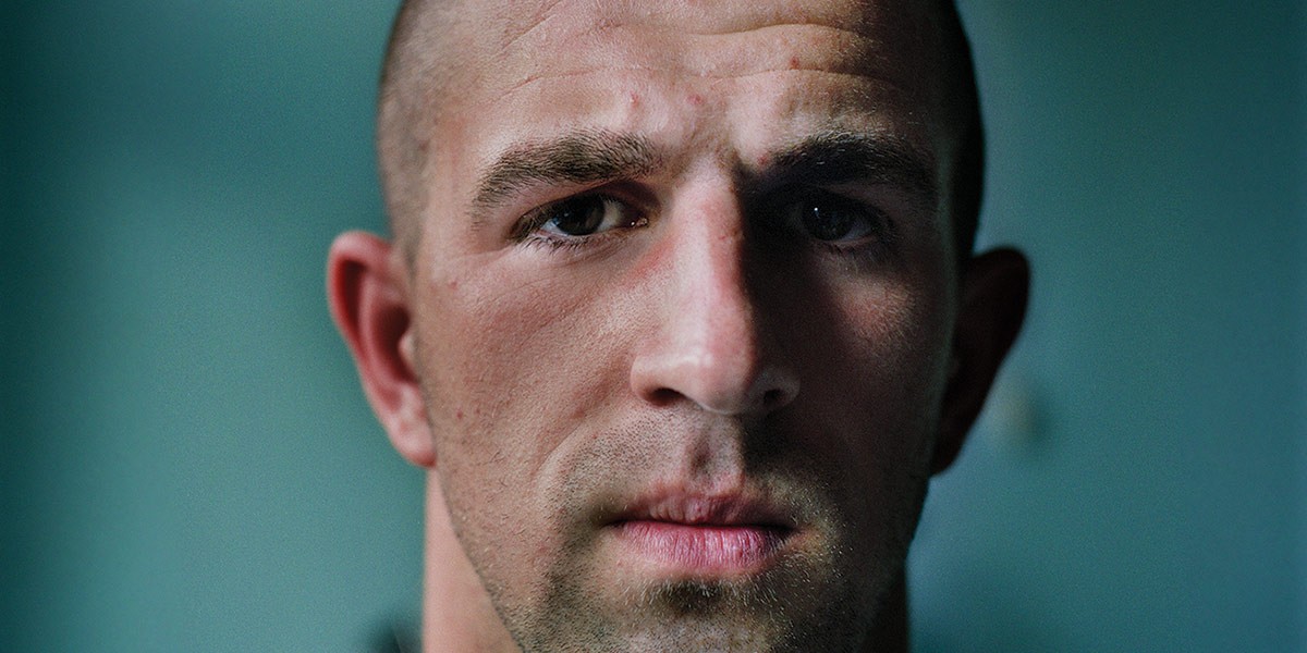 Close-up of a man's serious face with short hair against a teal blurred background.