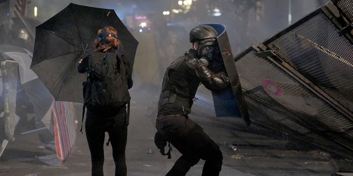 Protester with umbrella and gas mask next to a person in riot gear holding a shield near metal barricades at night.