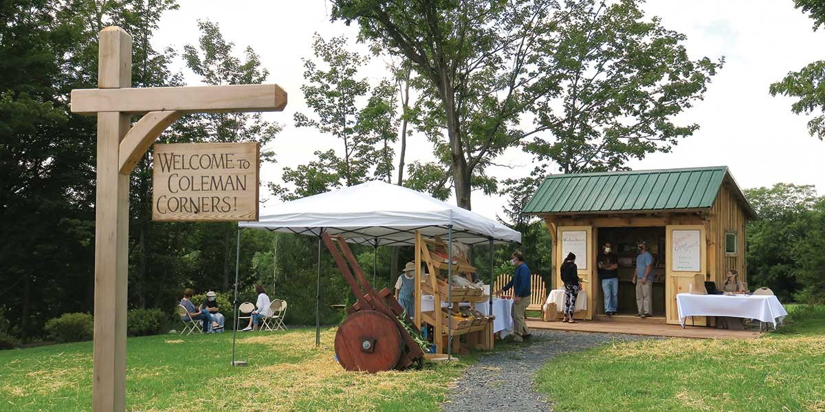 Outdoor market scene with wooden sign, booth under white canopy, small wooden building, and people talking nearby.