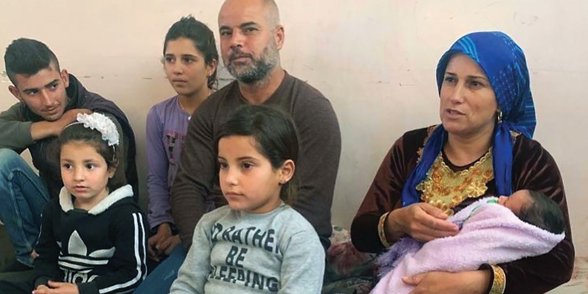Family of seven sitting together on the floor, including a woman holding a swaddled baby wrapped in a pink blanket.