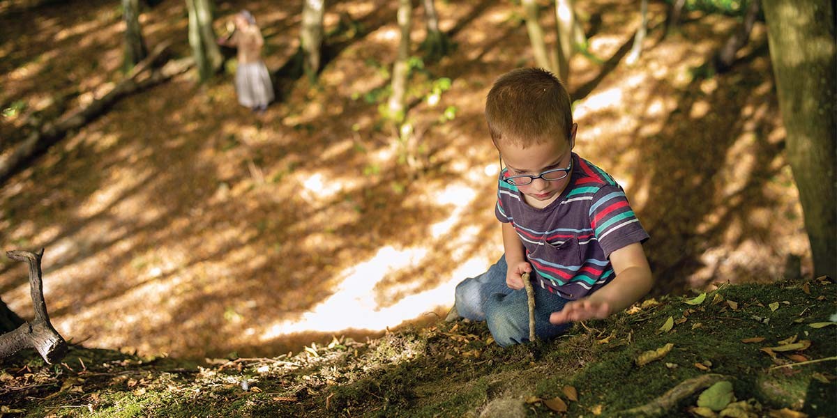 Boy with glasses climbing a mossy forest hill holding a stick in daylight.