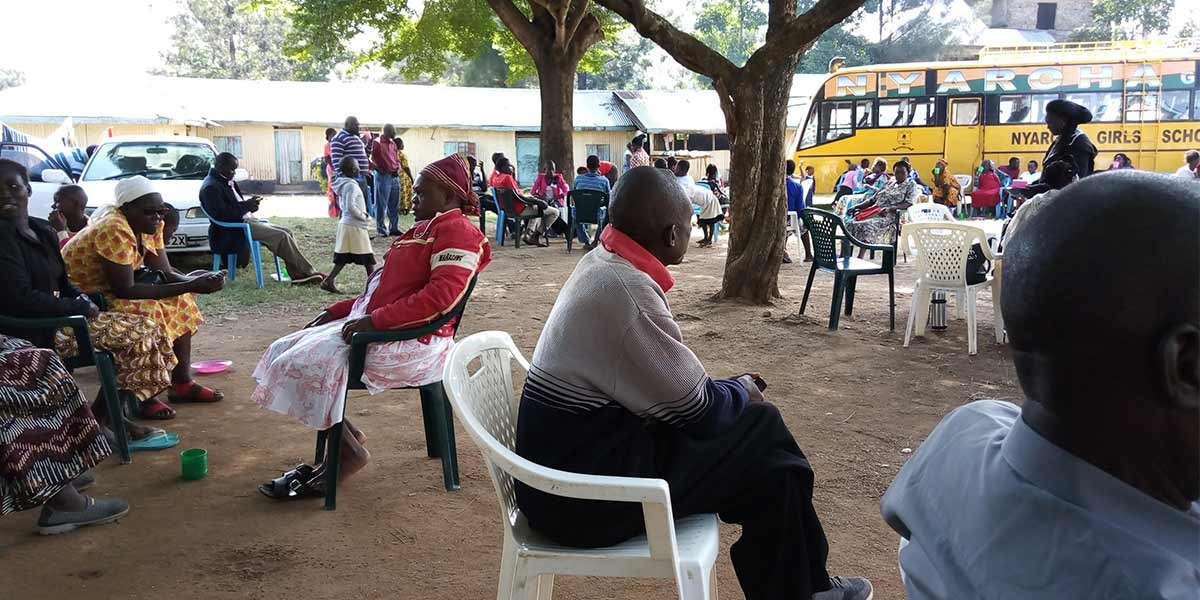 People sitting outdoors on plastic chairs under trees near a yellow school bus labeled Nyaroh Girls School.