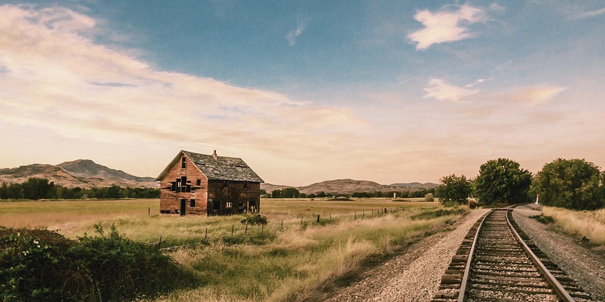 Abandoned wooden house in a grassy field beside curving railroad tracks and distant hills under a cloudy sky.