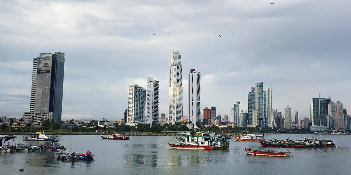 Boats floating on calm water with a city skyline of tall buildings and cloudy sky in the background.