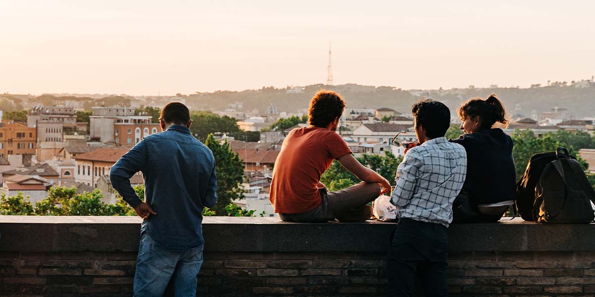 Four people sitting and standing on a stone wall overlooking a cityscape at sunset.