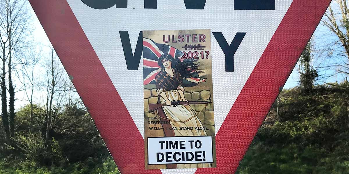 Sticker on road sign of a woman holding a rifle with UK flag, text says Ulster 1912 crossed out, 2021?, time to decide.