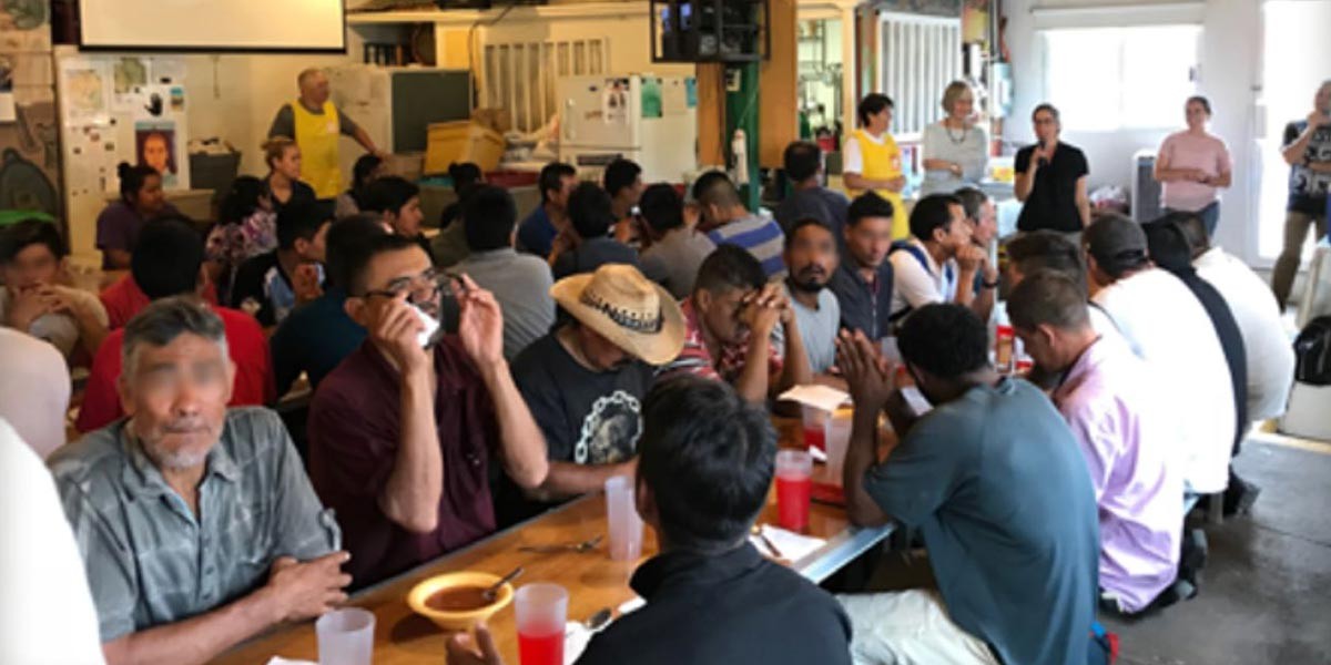 Group of people seated at tables eating and socializing in a casual indoor setting with volunteers standing.
