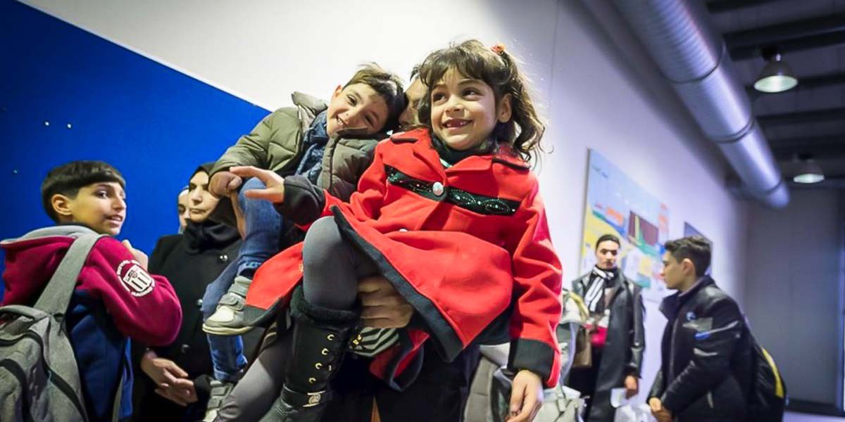 Group of children and adults indoors, with a smiling girl in a red coat held up in the center.