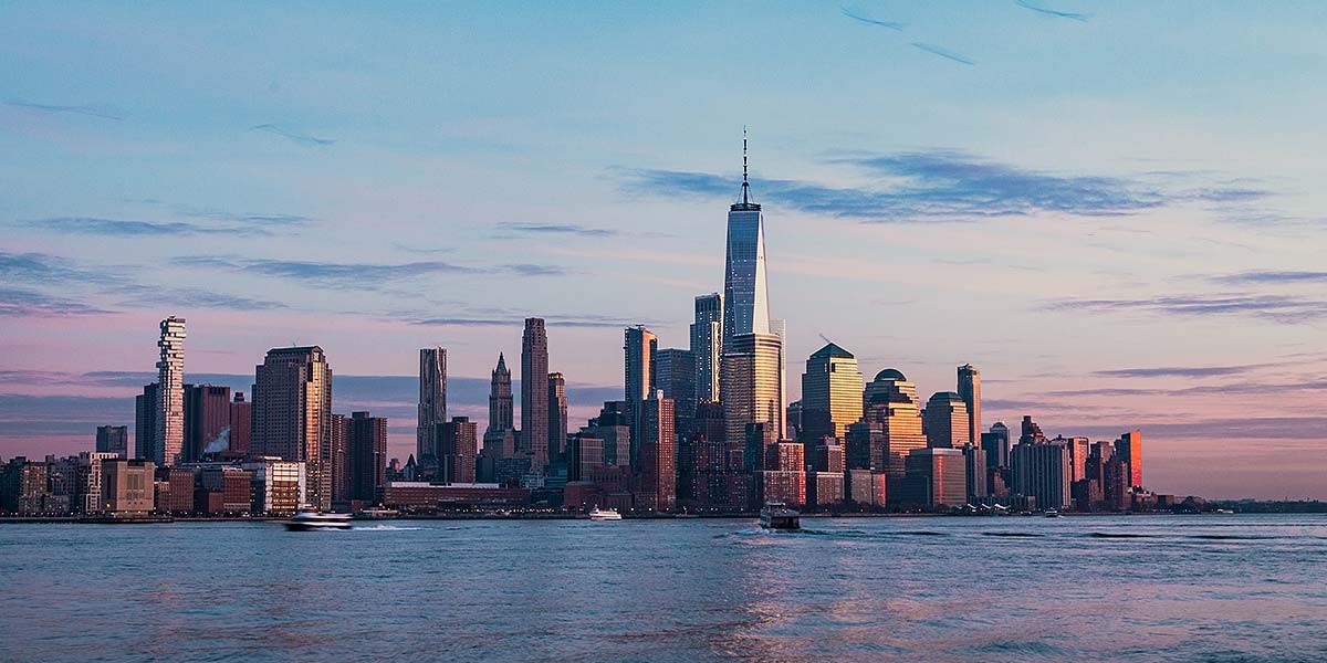 New York City skyline at sunset with One World Trade Center reflecting sunlight over water.
