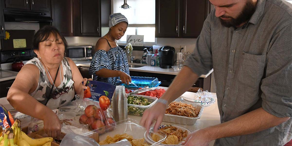 Three people serving food from trays on a kitchen counter with bananas and apples visible.