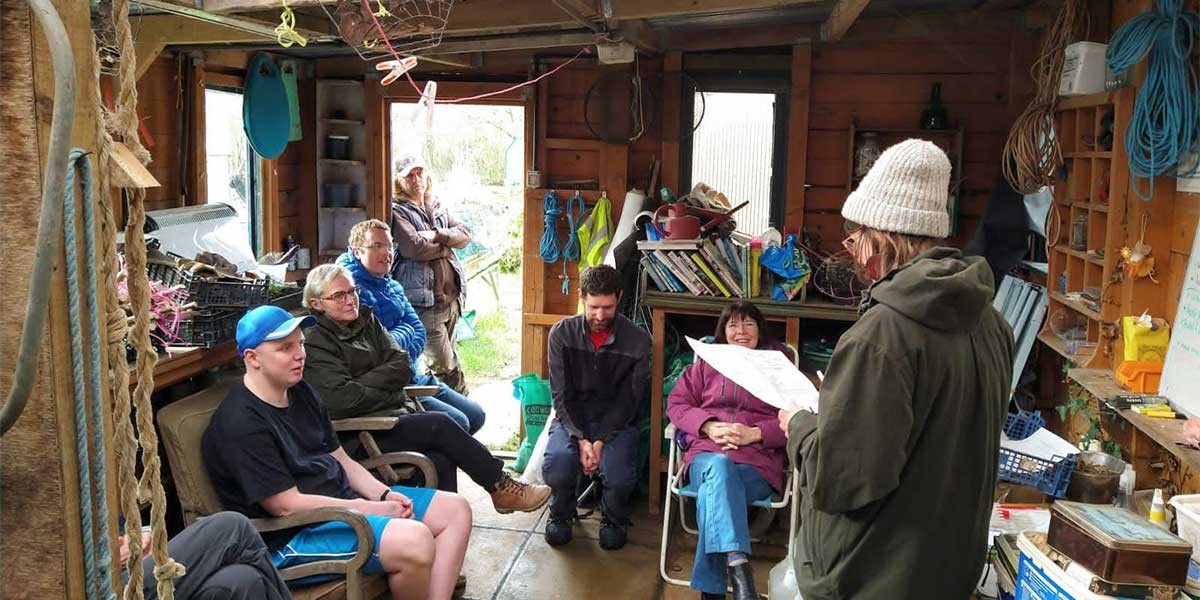 A group of people sitting and listening to a person reading from a paper in a rustic wooden room.