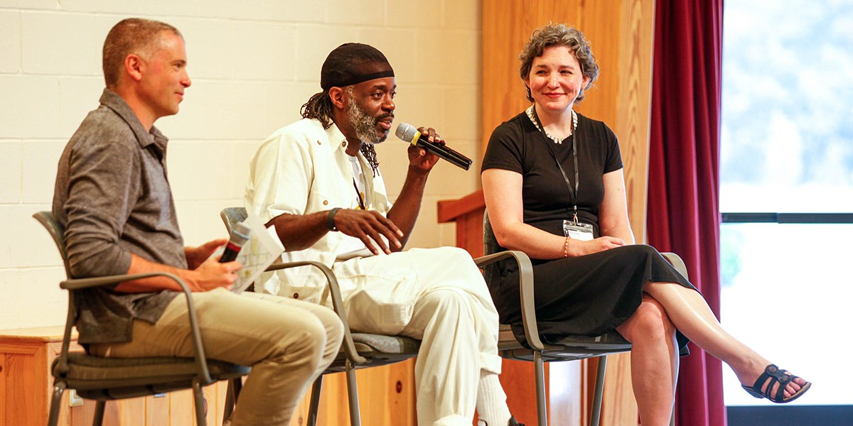 Three people sitting on chairs in conversation; the man in the middle is speaking into a microphone.