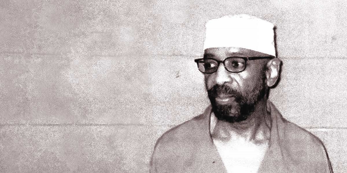 Black and white photo of a man with glasses, beard, and white cap against a plain wall.