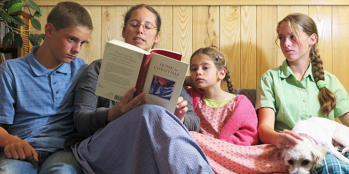 Woman reading a book titled 'Home for Christmas' to two girls and one boy seated on a couch with a dog.