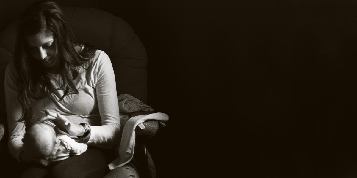 Mother sitting in chair gently cradling and looking at her baby in sepia-toned photo.