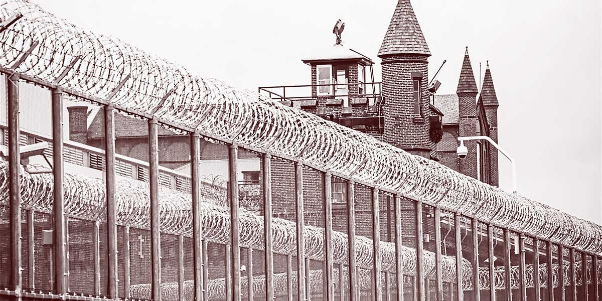Tall metal fence topped with coils of razor wire in front of an old brick prison with towers and a guard post.