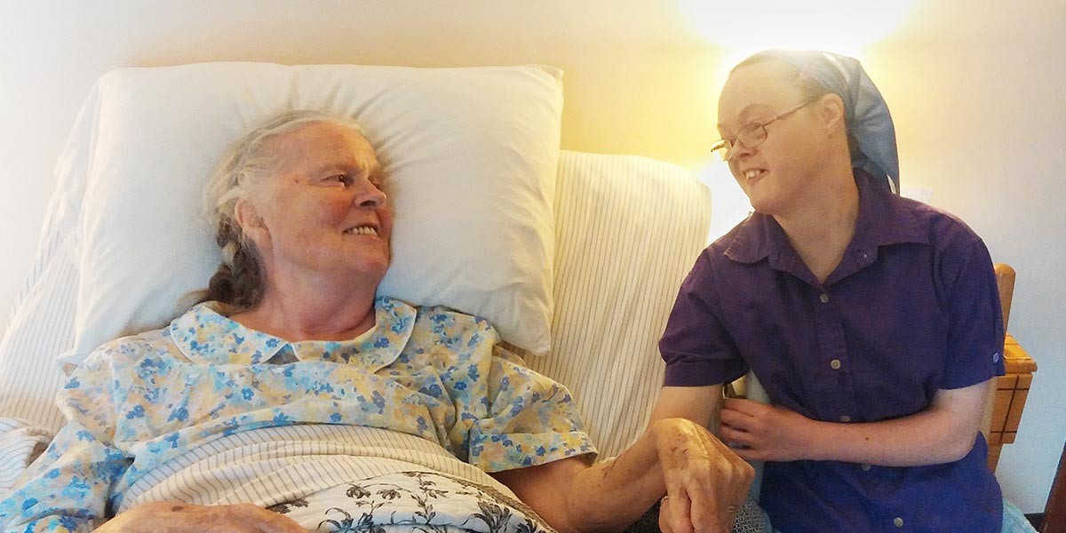 Elderly woman in bed holding hands and smiling at a younger woman wearing glasses and a headscarf.