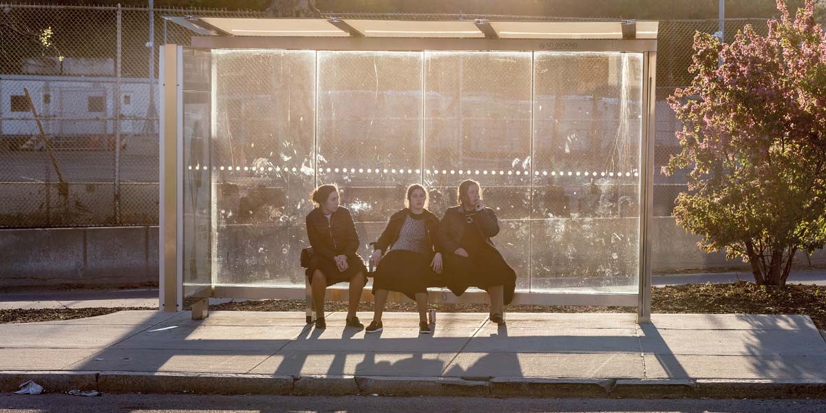 Three women dressed in dark clothes sitting on a bench inside a sunlit bus stop shelter.