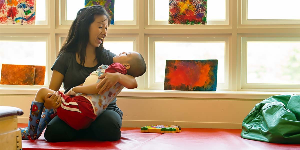 Woman joyfully holding a child wearing colorful socks on a red mat near bright windows with artwork.