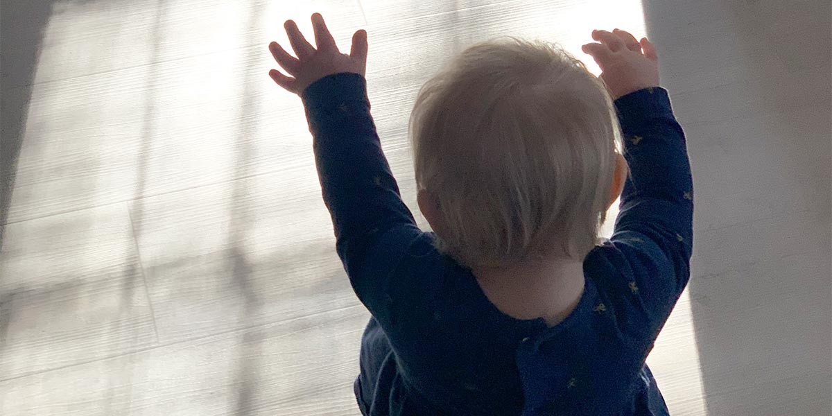 Baby with light hair raising arms sitting on a floor lit by sunlight through curtains.