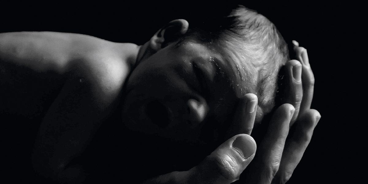Black and white photo of a newborn baby cradled gently in adult hands against a dark background.