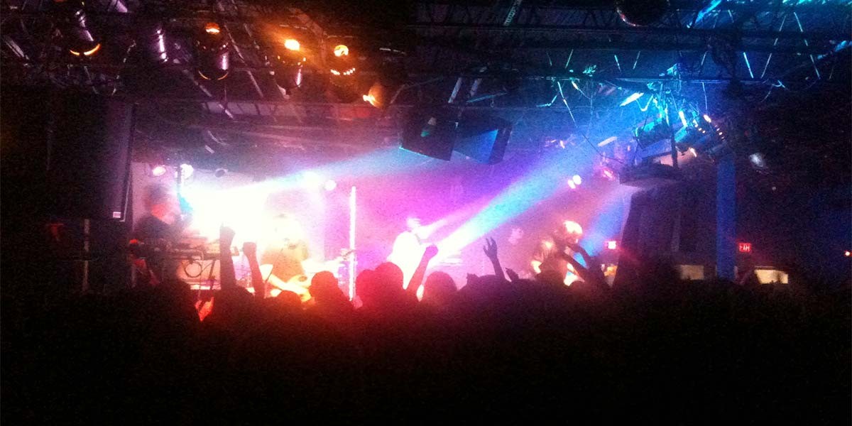 Silhouetted crowd with raised hands watching a brightly lit live music performance on stage.