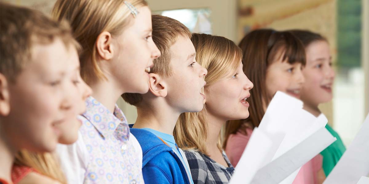 A group of children singing together while holding sheets of music in a bright room.