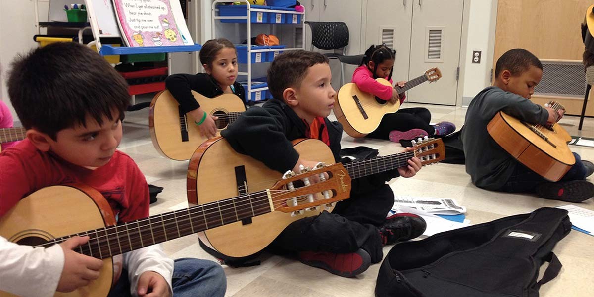 Children sitting on floor learning to play acoustic guitars in a classroom setting.