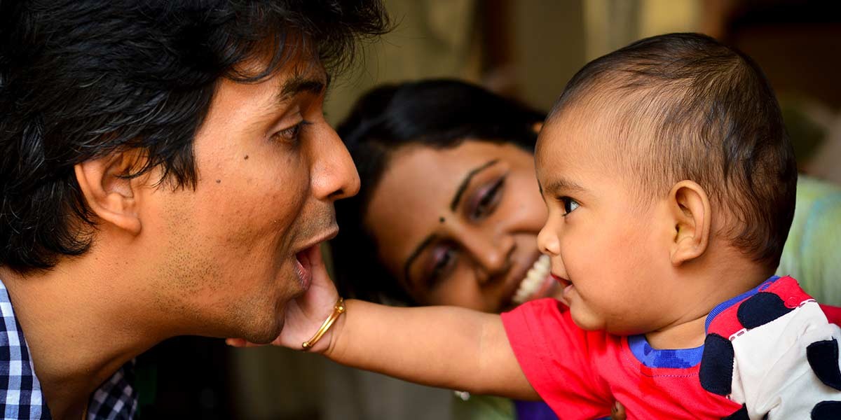 Baby in red shirt touching a man’s face while a woman smiles in the background.