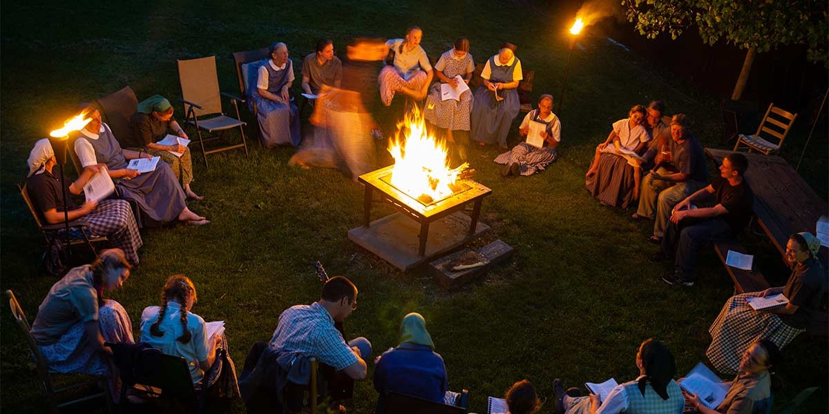 People in modest clothing sitting in a circle around a fire pit outdoors at night, reading and playing guitar.