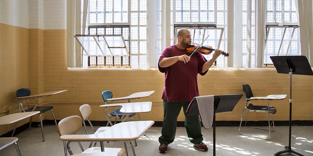 Man practicing violin alone in a brightly lit classroom with empty desks and music stands.