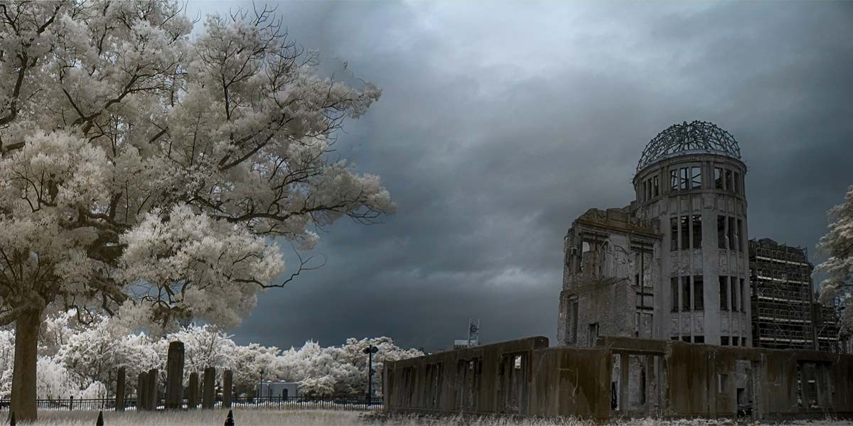 Ruined dome structure under dark sky beside large tree and fenced area with stone pillars.