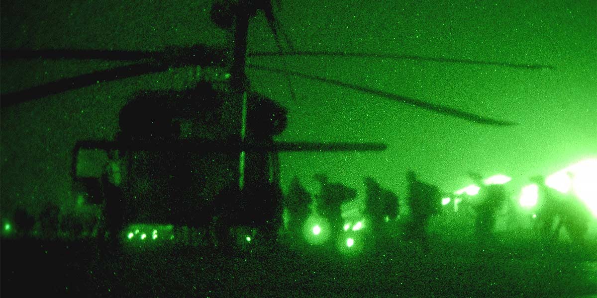 Night vision image of a military helicopter with soldiers disembarking in a green-lit scene.