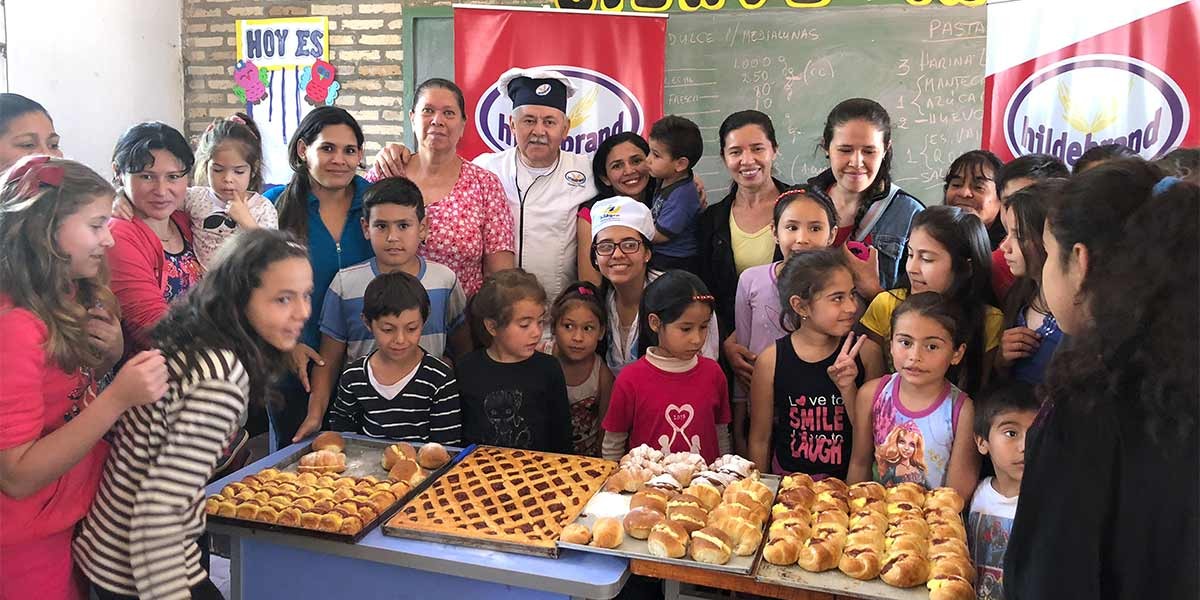 Group of children and adults standing behind a table with trays of assorted baked goods in a classroom.