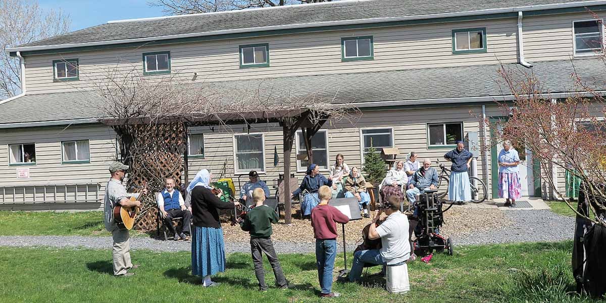 Group of people outdoors with children playing instruments and others seated near a building on a sunny day.