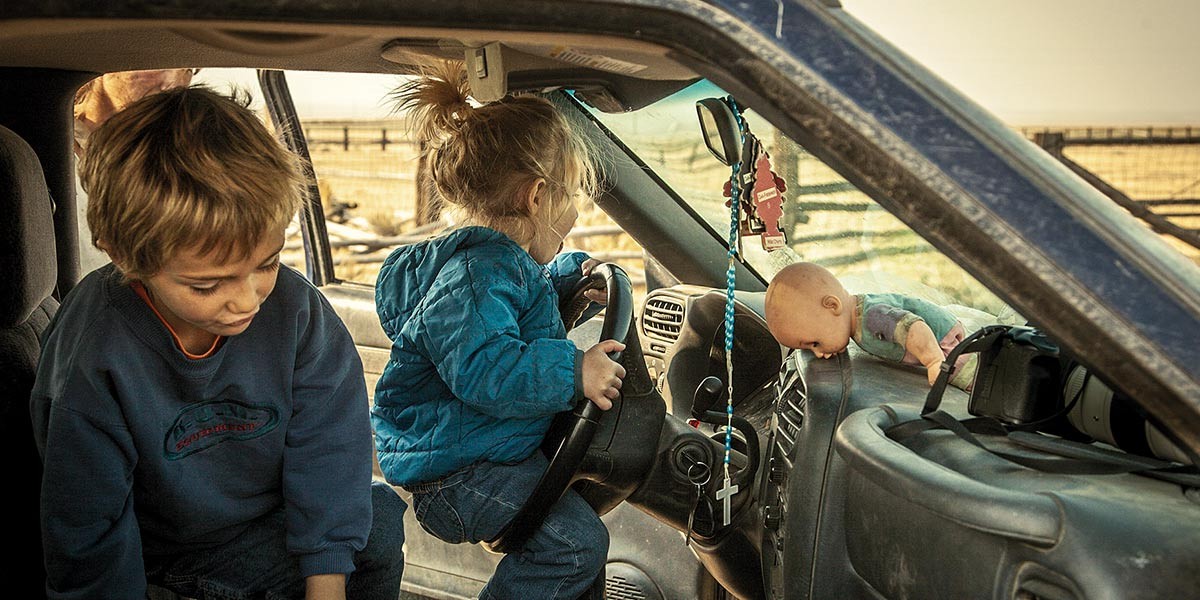 Two kids inside a vehicle: one boy sitting and one girl holding the steering wheel, with a doll on dashboard.