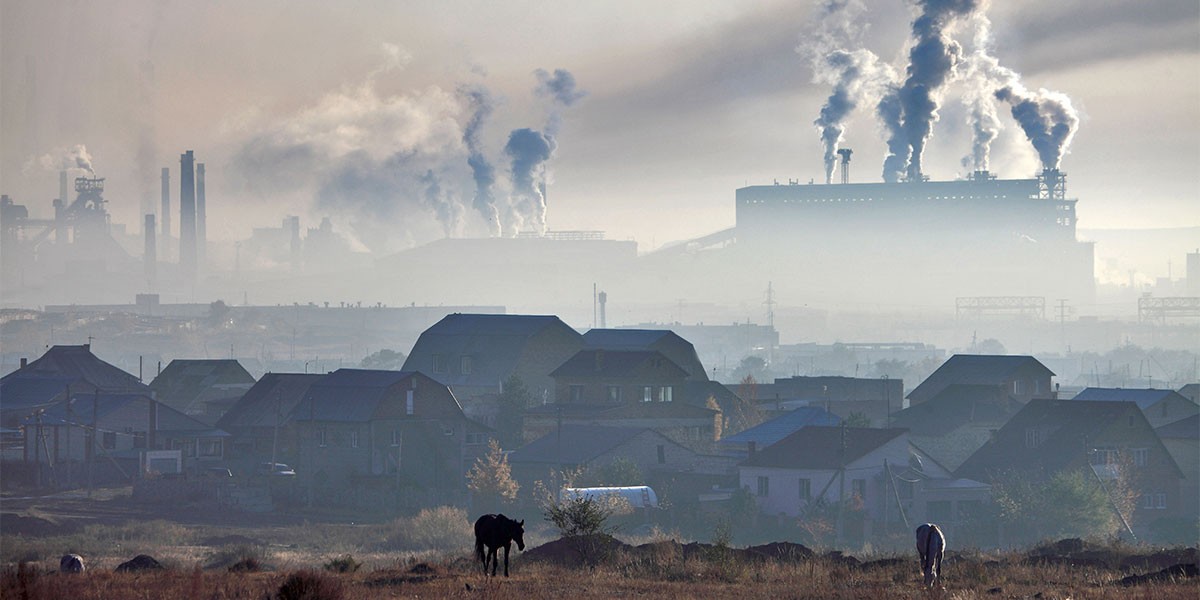 Industrial smokestacks emitting thick smoke over a rural village with houses and grazing horses.
