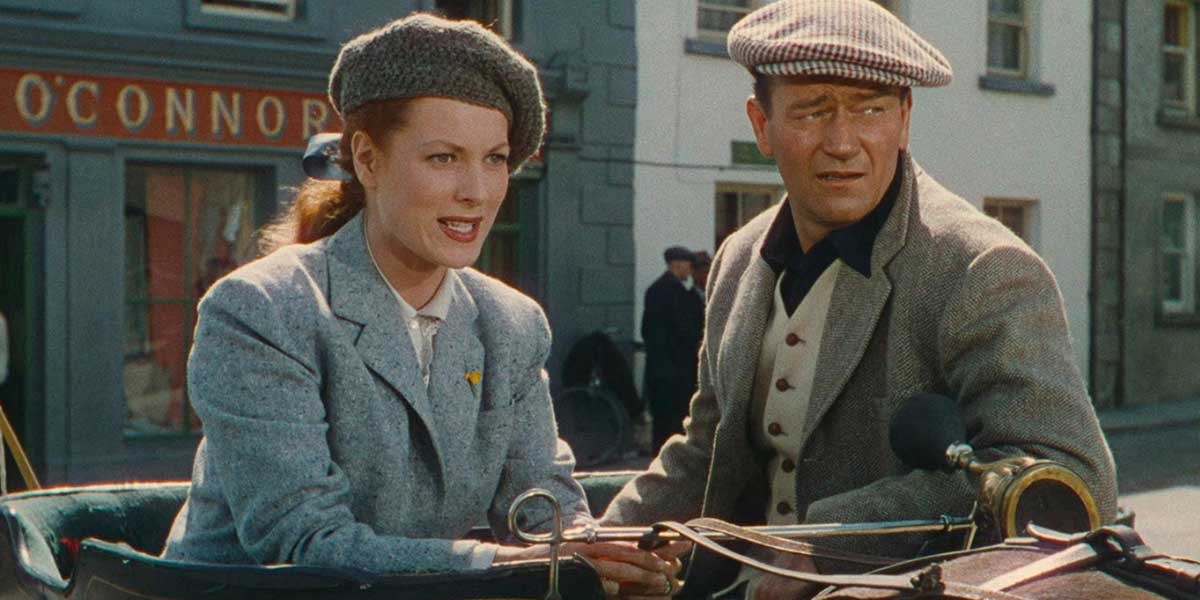 Man and woman in vintage attire sitting in an old-fashioned open vehicle on a street.