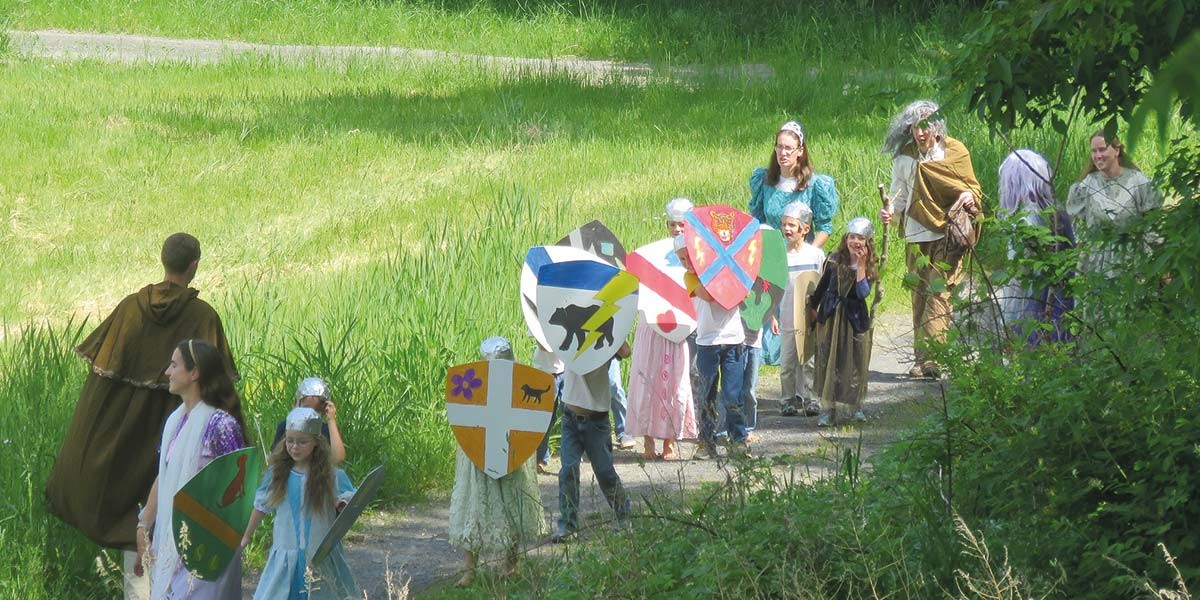 Group of children and adults in medieval costumes walking on a path with handmade shields in a green grassy area.