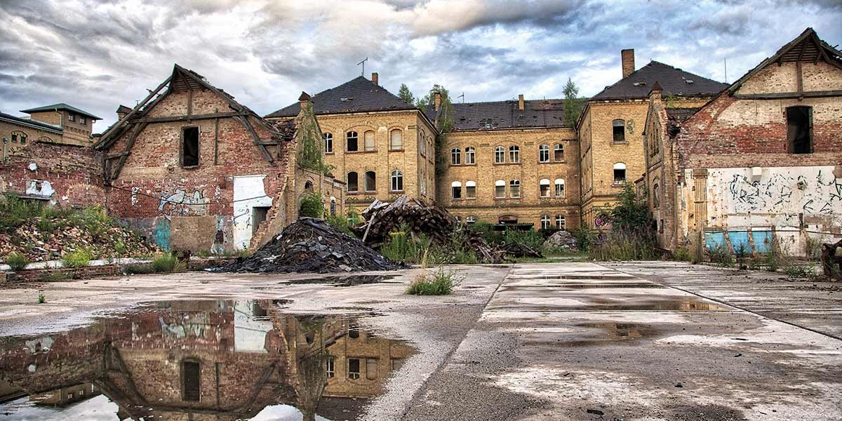 Abandoned brick buildings with broken roofs and scattered debris reflected in puddles on cracked concrete.