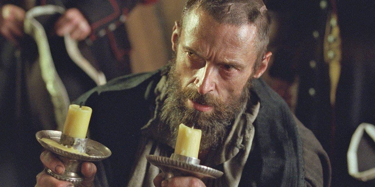 Bearded man with intense expression holding two candle holders with yellow candles.